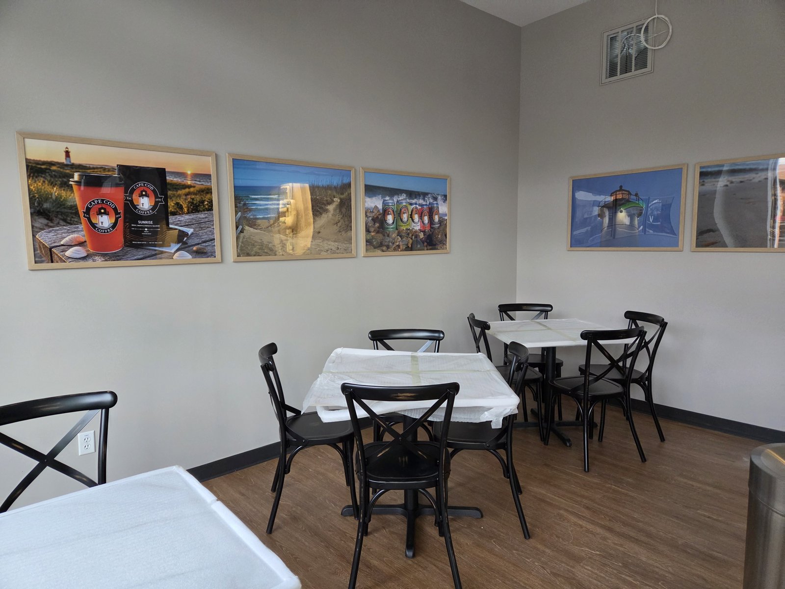 Black chairs and tables still wrapped in protective covering beneath framed photos of Cape Cod Coffee products, beaches, and lighthouses.  [Alt text by ChatGPT]